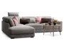 Moderne hjørnesofa betrukket med lysegråt stof, med en chaiselong på venstre side. Sofaen er pyntet med forskellige dekorative puder i rosa, brune og grå nuancer, som giver både komfort og stil. Foran sofaen står et rundt sofabord i træ med skrå ben, hvorpå der er en blomsterdekoration, stearinlys og små pyntegenstande. Sofaen har desuden et justerbart nakkestøtte for ekstra støtte.