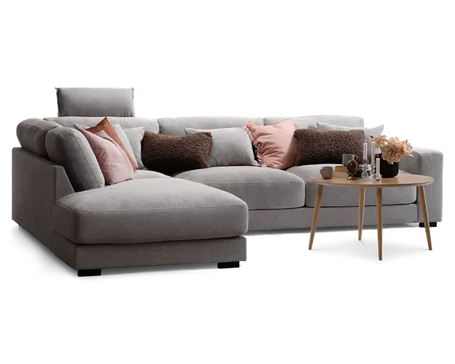 Moderne hjørnesofa betrukket med lysegråt stof, med en chaiselong på venstre side. Sofaen er pyntet med forskellige dekorative puder i rosa, brune og grå nuancer, som giver både komfort og stil. Foran sofaen står et rundt sofabord i træ med skrå ben, hvorpå der er en blomsterdekoration, stearinlys og små pyntegenstande. Sofaen har desuden et justerbart nakkestøtte for ekstra støtte.