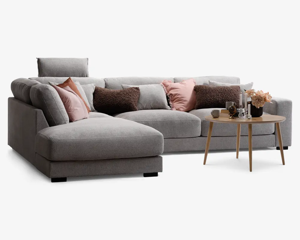 Moderne hjørnesofa betrukket med lysegråt stof, med en chaiselong på venstre side. Sofaen er pyntet med forskellige dekorative puder i rosa, brune og grå nuancer, som giver både komfort og stil. Foran sofaen står et rundt sofabord i træ med skrå ben, hvorpå der er en blomsterdekoration, stearinlys og små pyntegenstande. Sofaen har desuden et justerbart nakkestøtte for ekstra støtte.