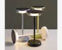 Tre moderne bordlamper med minimalistisk design, der har flade, runde toppe med integreret LED-belysning langs kanten. Lamperne har slanke, cylindriske stænger og cirkulære baser, vist i sort, hvid og lysegrøn. Toppen har en tekstureret overflade, muligvis til touch-betjening eller som dekorativ detalje. Lamperne står oprejst, og én ligger vandret, hvilket fremhæver den stabile base og det elegante udtryk.