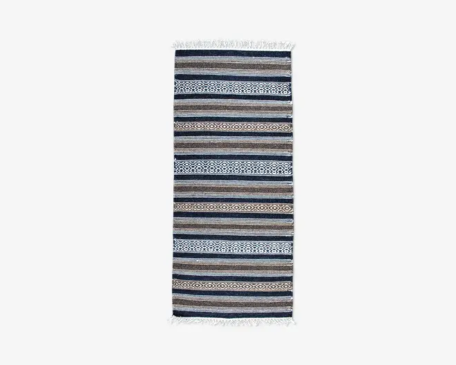 Rektangulært tekstil med skiftende horisontale striber i blå, brune, beige og hvide nuancer. Nogle striber har indviklede geometriske mønstre, herunder diamant- og zigzagmønstre. Enderne er afsluttet med korte hvide frynser.