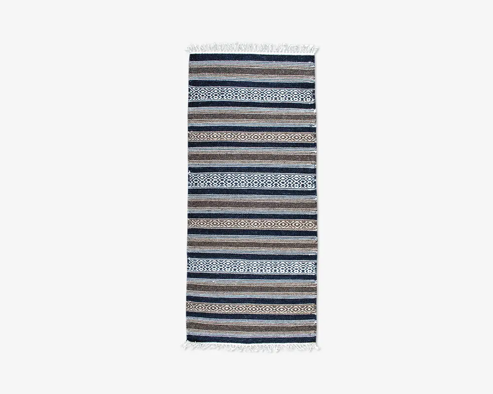 Rektangulært tekstil med skiftende horisontale striber i blå, brune, beige og hvide nuancer. Nogle striber har indviklede geometriske mønstre, herunder diamant- og zigzagmønstre. Enderne er afsluttet med korte hvide frynser.