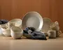 Et koordineret sæt keramikservice med mat beige og cremefarvet gradient. Sættet indeholder store middagstallerkener, mindre sidetallerkener, dybe skåle og krus, alle med en diskret plettet tekstur og blødt afrundede kanter. Krusene har brede, komfortable hanke, og skålene er dybe med en glat, blank inderside. Det overordnede design er moderne, men rustikt, med et håndlavet udtryk. Mørkegrå stofservietter er lagt over nogle af delene og giver et flot farvespil.