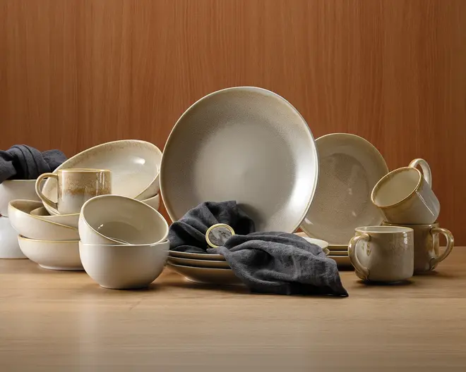 Et koordineret sæt keramikservice med mat beige og cremefarvet gradient. Sættet indeholder store middagstallerkener, mindre sidetallerkener, dybe skåle og krus, alle med en diskret plettet tekstur og blødt afrundede kanter. Krusene har brede, komfortable hanke, og skålene er dybe med en glat, blank inderside. Det overordnede design er moderne, men rustikt, med et håndlavet udtryk. Mørkegrå stofservietter er lagt over nogle af delene og giver et flot farvespil.