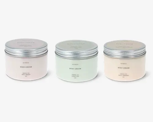Tre runde krukker med brede, flade kroppe og metalliske skruelåg. Hver krukke har en forskellig pastelfarve: sart lyserød, lysegrøn og lys beige. De minimalistiske etiketter har enkel, elegant tekst, der angiver produkttype og duft, med mærkenavnet centreret øverst. Krukkerne har et rent, moderne design, og lågene har en diskret rillet struktur for bedre greb.