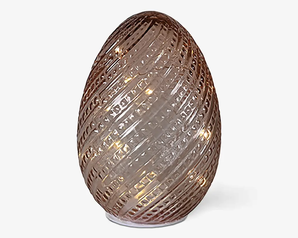 Dekorativt objekt formet som et æg, lavet af gennemsigtigt glas med et tekstureret, spiralformet mønster. Glasset har en let metallisk eller reflekterende overflade, og små varme hvide LED-lys er synlige indeni, hvilket skaber en blød, glødende effekt. Overfladen har forhøjede riller og udskæringer, der forstærker lysspredningen.