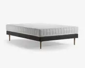 Sengen har et moderne, minimalistisk design med en tyk, quiltet madras i hvid, som har en tekstureret, mønstret overflade for ekstra komfort. Bunden er betrukket med et mørkt stof, hvilket giver en elegant kontrast til madrassen. Sengen står på fire koniske træben, hvilket giver et solidt og stilfuldt udtryk samt nem rengøring under sengen.
