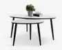 Sæt med to indskudsborde med runde, hvide bordplader og slanke, sorte skrå ben. Det større bord er dekoreret med en potteplante, et blåt krus, to glas og en stak bøger. Det mindre bord kan skubbes delvist ind under det større, hvilket giver fleksibel indretning og pladsbesparende opbevaring. Designet er moderne og minimalistisk.