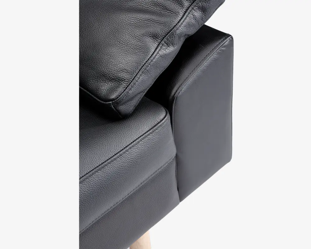 Nærbillede af et sofahjørne med sort tekstureret læderbetræk, en blød sædepude, en matchende ryghynde og en polstret armlæn. Sofaen har lyse træben, som kan ses nederst.