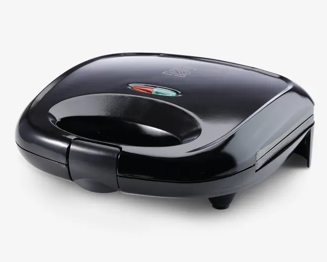 En kompakt, sort maskine med buet top og integreret håndtag for nem åbning. På toppen er der to indikatorlamper – en rød og en grøn – som viser strøm- og klarstatus. Apparatet har en sikker lås foran og står på fire små fødder for stabilitet. Den har en glat, blank overflade, der giver et moderne udseende.