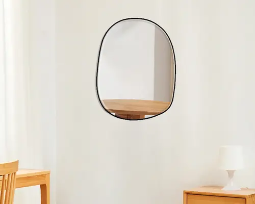 Vægmonteret spejl med uregelmæssig, afrundet form og en tynd sort ramme. Spejlet har et glat og moderne udtryk og reflekterer en del af et træbord. Designet er minimalistisk og passer godt til moderne indretning.