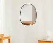Vægmonteret spejl med uregelmæssig, afrundet form og en tynd sort ramme. Spejlet har et glat og moderne udtryk og reflekterer en del af et træbord. Designet er minimalistisk og passer godt til moderne indretning.