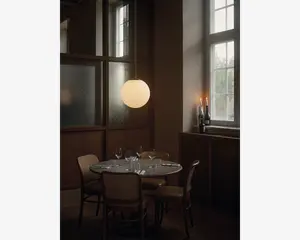 Et rundt spisebord dækket til fire personer med stole med flet-ryg, placeret i et blødt oplyst rum. Bordet er dækket med klare vinglas, vandglas, hvide tallerkener og sølvbestik, samt hvide foldede servietter ved hver plads. Over bordet hænger en stor, kugleformet lampe i matteret glas, der udsender et varmt lys. I baggrunden er der høje vinduer med naturligt lys og et sidebord med to tændte stearinlys og vinflasker, hvilket skaber en hyggelig og elegant stemning.