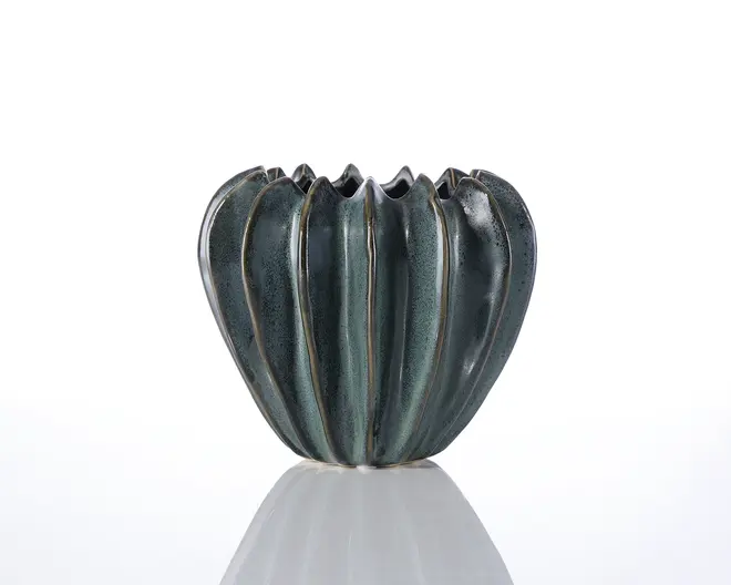 Et keramikkar med en blank, mørkegrøn glasur og en unik, skulpturel form, der minder om et græskar eller en squash. Overfladen har lodrette riller og fordybninger, og kanten er ujævn og bølget, hvilket giver et organisk udtryk. Glasuren har diskrete farvevariationer, som understreger det håndlavede præg.