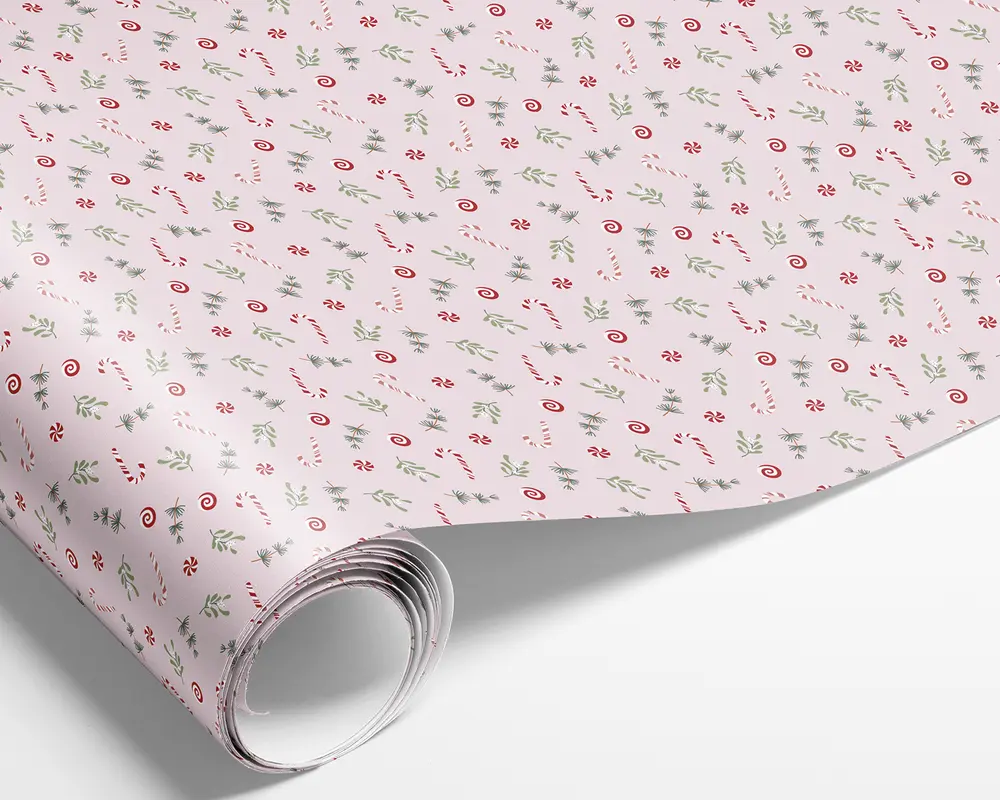 Billedet viser en rulle papir med et festligt mønster. Baggrunden er en blød pastelpink, og designet har små illustrationer af rød- og hvidstribede slikstokke, pebermyntebolsjer og grønne grene af kristtorn og fyr. Mønstret er jævnt fordelt og gentages over hele overfladen, hvilket giver et muntert og julet udtryk.