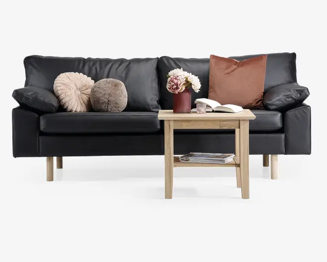 En moderne sort lædersofa med træben, udstyret med tre dekorative pyntepuder i beige, brun og taupe. Foran sofaen står et lyst sofabord i træ med en hylde under, hvor der ligger magasiner. På bordpladen står en rød vase med hvide og lyserøde blomster, et glas og en åben bog.