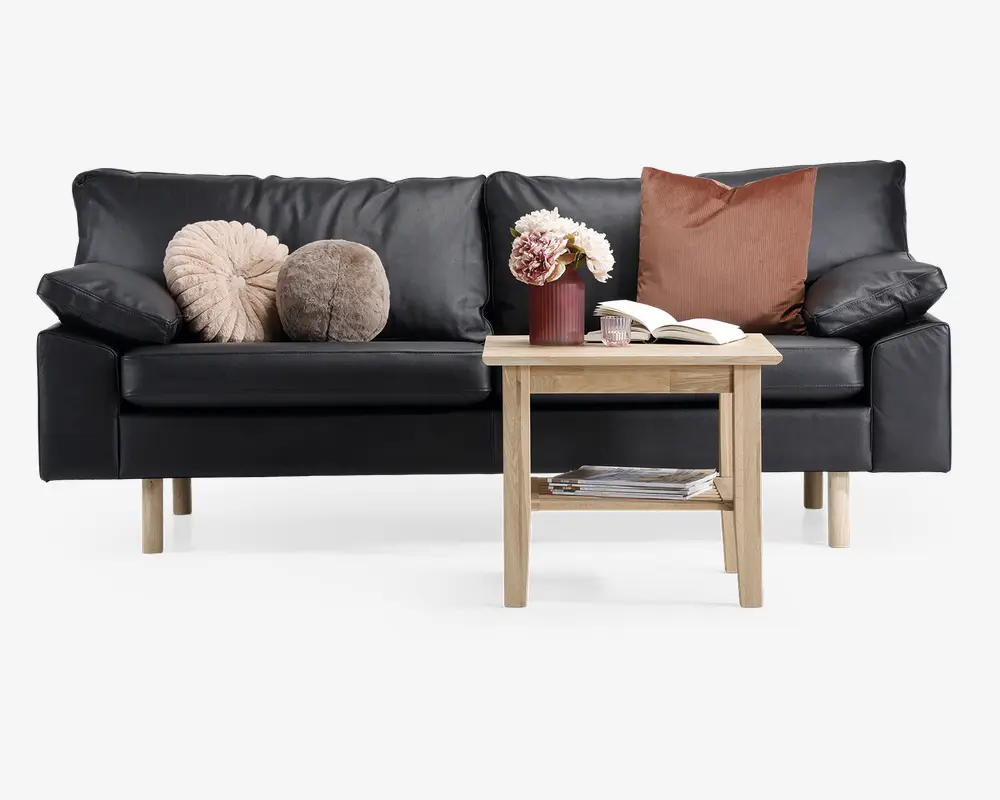 En moderne sort lædersofa med træben, udstyret med tre dekorative pyntepuder i beige, brun og taupe. Foran sofaen står et lyst sofabord i træ med en hylde under, hvor der ligger magasiner. På bordpladen står en rød vase med hvide og lyserøde blomster, et glas og en åben bog.
