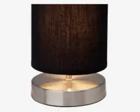 En lampe med en cylindrisk sort stofskærm og en rund, børstet metalfod. Foden har en glat, reflekterende overflade, og lampen udsender et varmt, blødt lys under skærmen. Designet er moderne og minimalistisk.
