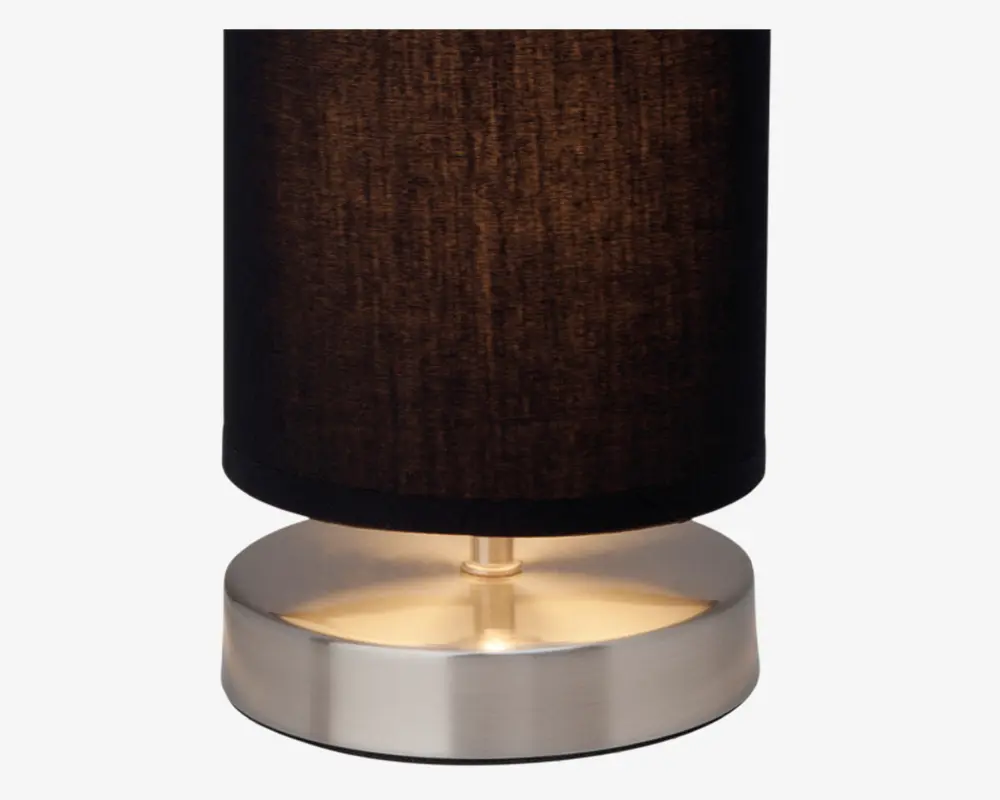 En lampe med en cylindrisk sort stofskærm og en rund, børstet metalfod. Foden har en glat, reflekterende overflade, og lampen udsender et varmt, blødt lys under skærmen. Designet er moderne og minimalistisk.