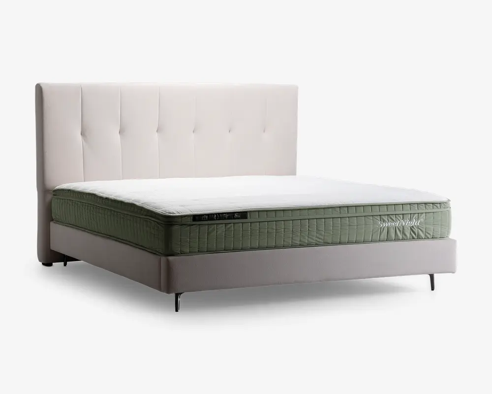 Billedet viser en seng med et højt, polstret sengegærde i lysebeige farve med lodrette, tuftede detaljer. Madrassen har et grønt, quiltet stofbetræk med et synligt mærkelabel på siden, og en hvid topmadras giver ekstra komfort. Sengerammen er betrukket med samme lysebeige stof og står på slanke, metalliske ben, hvilket giver et moderne og elegant udtryk.