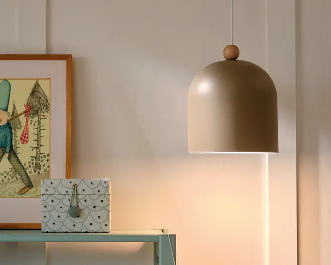 En mat beige pendellampe med kuppelformet skærm og en lille trædetalje øverst, der kaster et varmt lys på væggen.