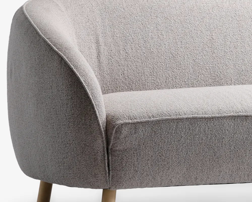 Nærbillede af en stol med lysegråt tekstureret stofbetræk, afrundet armlæn og ryglæn samt lyse træben. Stoffet ser blødt og slidstærkt ud, og stolens form er moderne og indbydende.