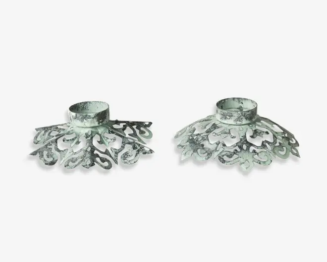 Sæt med to metalholdere med et udsmykket, vintage-inspireret design. Hver holder har en slidt, patineret overflade i sølvfarvet nuance og detaljerede udskæringer, der danner en dekorativ, bred fod. Toppen har en rund åbning, der passer til et lys eller lignende genstand. Udtrykket er antikt og elegant, perfekt til at tilføje klassisk charme til indretningen.