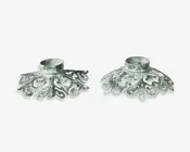 Sæt med to metalholdere med et udsmykket, vintage-inspireret design. Hver holder har en slidt, patineret overflade i sølvfarvet nuance og detaljerede udskæringer, der danner en dekorativ, bred fod. Toppen har en rund åbning, der passer til et lys eller lignende genstand. Udtrykket er antikt og elegant, perfekt til at tilføje klassisk charme til indretningen.