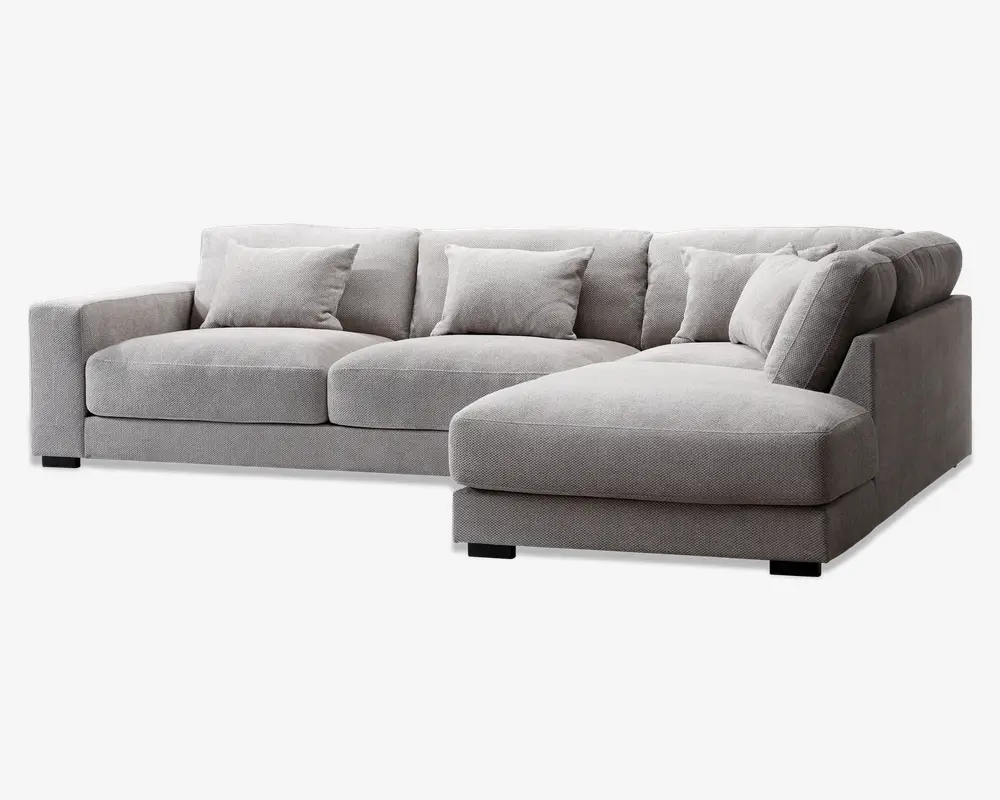 Moderne hjørnesofa i lysegråt stof med chaiselong til højre. Sofaen har brede, lavprofilerede armlæn og bløde, dybe sædehynder for ekstra komfort. Leveres med fem matchende ryghynder for øget støtte. Betrækket har en blød, tekstureret overflade, og sofaen står på korte, solide sorte ben.