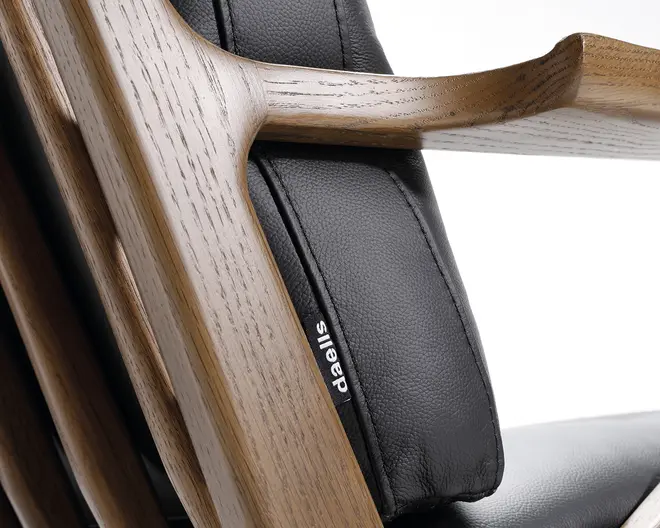 Nærbillede af en stol med en glat, lys træramme med naturligt åremønster. Stolen har sorte, teksturerede læderhynder til sæde og ryg, hvilket giver et moderne og elegant udtryk. Syningerne på hynderne er tydelige, og der er et lille stofmærke med mærket 'daells' fastgjort på siden af rygpuden.