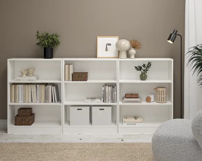 Et moderne, hvidt reolsystem med tre sektioner, rene linjer og åbne hylder. Hylderne er dekoreret med en blanding af bøger, dekorative genstande som vaser, kurve og indrammet kunst. Nogle hylder har opbevaringskasser, mens andre viser små skulpturer og planter. Reolen står på et lyst trægulv op ad en taupefarvet væg, med en sort gulvlampe og en hyggelig lænestol i nærheden, hvilket skaber et minimalistisk og organiseret udtryk.