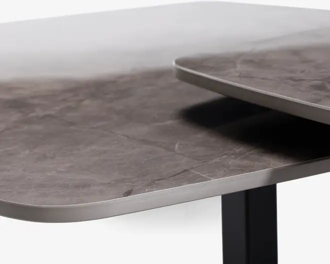 Nærbillede af en bordplade med en glat, mat stenlignende overflade i mørkegrå farve. Kanterne er afrundede og afsluttet med en tynd metallisk kant, hvilket giver et moderne og elegant udtryk. Bordet ser ud til at have et lagdelt eller udtrækkeligt design, da en anden overflade ses lidt over hovedpladen.