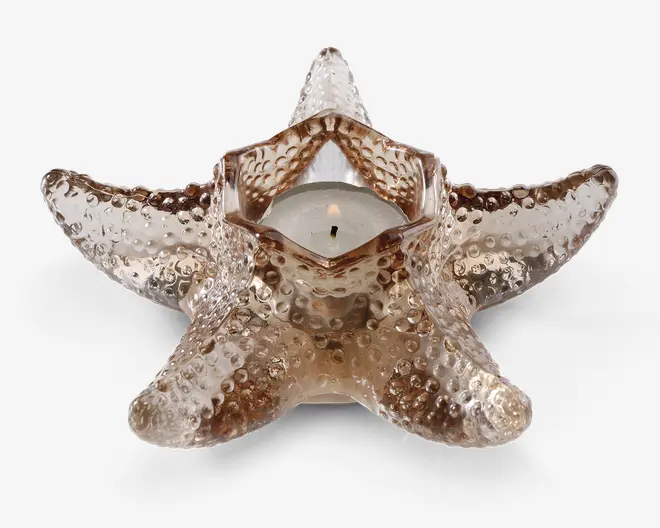 En dekorativ lysestage formet som en søstjerne, lavet af tekstureret glas med en gennemsigtig, lysebrun nuance. Lysestagen har et centralt hulrum, der passer til et lille fyrfadslys, og overfladen er dækket af forhøjede knopper, der efterligner en søstjernes naturlige udseende.
