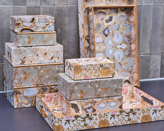 Et udvalg af dekorative opbevaringsæsker og bakker med indviklede mønstre. Overfladerne er udsmykket med motiver af fugle, blomster og grene i guld, beige og pastelfarver samt felter, der ligner poleret agatsten. Æskerne er stablet i forskellige størrelser, og bakkerne har udskårne håndtag for nem transport. Det samlede udtryk er elegant og udsmykket, velegnet til stilfuld hjemmeopbevaring eller som gave.