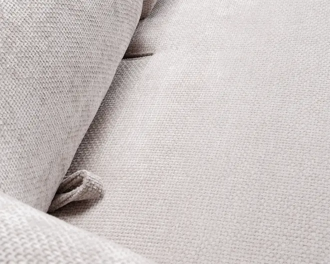 Nærbillede af et lysebeige, tekstureret stof med et diskret vævet mønster. Overfladen ser blød og polstret ud med synlige knapdetaljer og et indbydende, behageligt udtryk. Stoffet har en let forhøjet struktur, som øger den taktile fornemmelse og komforten.