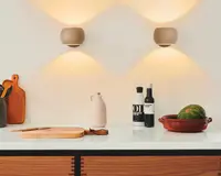 En moderne køkkenbordplade med en ren, hvid overflade. Til venstre står to skærebrætter af træ, det ene oprejst og det andet fladt, med en kniv liggende på det flade bræt. En sort beholder indeholder forskellige køkkenredskaber. I midten står en lyserød keramikkande. Til højre står to flasker, den ene mærket 'OIL' og den anden en mørk flaske med hvid etiket. Helt til højre ligger et rundt, grønt græskar i et rødt keramisk fad. Baggrunden er en hvid væg med to beige væglamper, der udsender varmt, stemningsfuldt lys både opad og nedad.