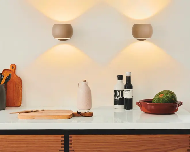 En moderne køkkenbordplade med en ren, hvid overflade. Til venstre står to skærebrætter af træ, det ene oprejst og det andet fladt, med en kniv liggende på det flade bræt. En sort beholder indeholder forskellige køkkenredskaber. I midten står en lyserød keramikkande. Til højre står to flasker, den ene mærket 'OIL' og den anden en mørk flaske med hvid etiket. Helt til højre ligger et rundt, grønt græskar i et rødt keramisk fad. Baggrunden er en hvid væg med to beige væglamper, der udsender varmt, stemningsfuldt lys både opad og nedad.