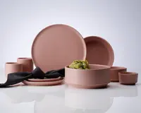 Et koordineret sæt af matte, støvet rosa keramikstel, bestående af store og små tallerkener, skåle og kopper. Delene har et minimalistisk, moderne design med glatte, udekorerede overflader og afrundede kanter. Sættet er arrangeret på en reflekterende overflade, med en sort stofserviet lagt over to stablede tallerkener og et artiskokhoved placeret i en af skålene. Udtrykket er elegant og nutidigt.
