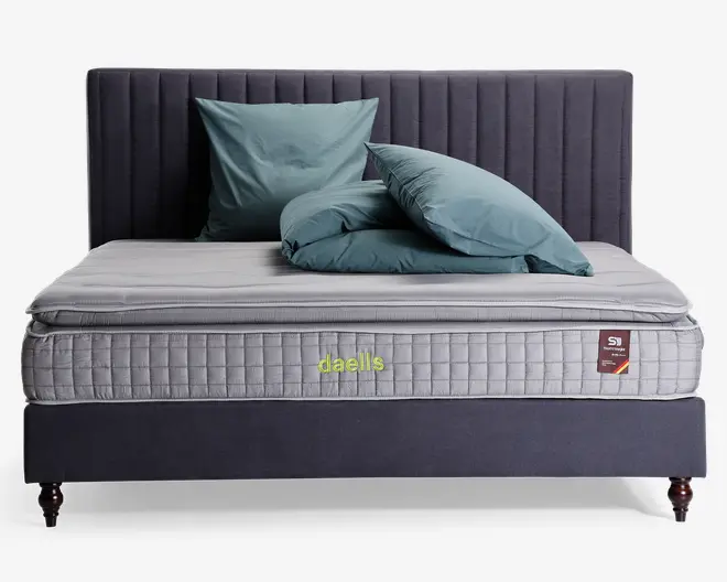 En moderne seng med en høj, lodret-stiksyet sengegavl betrukket med mørkegråt stof. Madrassen er tyk og har et quiltet, lysegråt betræk med et synligt 'daells'-logo broderet i gulgrøn. Sengen er redt med blågrønne puder og en matchende dyne, der er pænt arrangeret. Sengerammen har korte, mørke træben, hvilket giver et solidt og elegant udtryk.