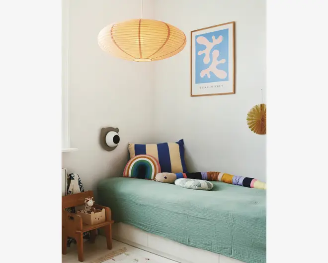Et hyggeligt hjørne af et børneværelse med en enkeltseng, der har et lysegrønt, tekstureret sengetæppe. Sengen er pyntet med sjove puder, herunder en stor blå og gulstribet pude, en regnbueformet pude og en lang, flerfarvet pøllepude. En lille bamse ligger på sengen. Over sengen hænger en rund papirlampe, der giver blødt lys. På væggen er der et indrammet abstrakt kunsttryk i blå og hvid. Ved siden af sengen står en lille træstol med en bamse og en kurv, hvilket giver rummet varme og charme.