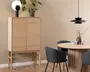 Høj skab i lyst træ med to lodrette rillede låger, stående på slanke ben. Skabet har et minimalistisk design med rene linjer og en naturlig finish. Ovenpå står en moderne bordlampe med en rund hvid kugle og sort base samt en grøn potteplante. Skabet er placeret op ad en beige væg og står på et trægulv.