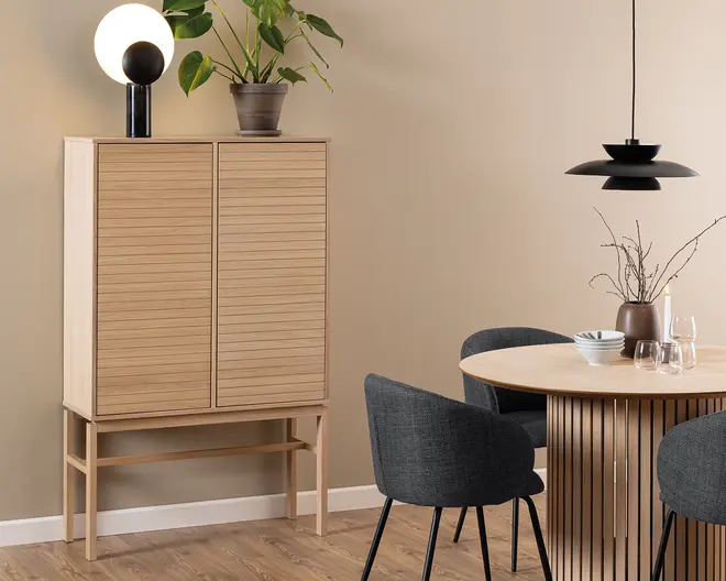 Høj skab i lyst træ med to lodrette rillede låger, stående på slanke ben. Skabet har et minimalistisk design med rene linjer og en naturlig finish. Ovenpå står en moderne bordlampe med en rund hvid kugle og sort base samt en grøn potteplante. Skabet er placeret op ad en beige væg og står på et trægulv.