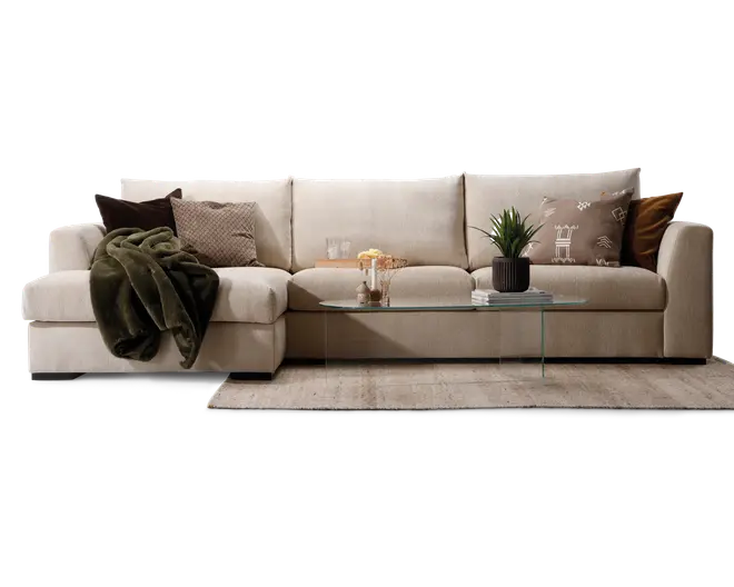 Rummelig L-formet sofa betrukket med lysebeige stof, udstyret med brede, bløde sæde- og ryghynder for ekstra komfort. Sofaen er pyntet med forskellige pyntepuder i brune, beige og mønstrede designs samt et mørkegrønt tæppe lagt over den ene armlæn. Foran sofaen står et minimalistisk sofabord i glas med en potteplante, bøger og dekorative genstande. Opsætningen står på et neutralt farvet tæppe, hvilket skaber en hyggelig og indbydende stue.