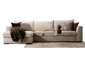 Rummelig L-formet sofa betrukket med lysebeige stof, udstyret med brede, bløde sæde- og ryghynder for ekstra komfort. Sofaen er pyntet med forskellige pyntepuder i brune, beige og mønstrede designs samt et mørkegrønt tæppe lagt over den ene armlæn. Foran sofaen står et minimalistisk sofabord i glas med en potteplante, bøger og dekorative genstande. Opsætningen står på et neutralt farvet tæppe, hvilket skaber en hyggelig og indbydende stue.