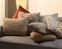 En opbygget samling pyntepuder på en grå sofa med beige og brune stribede puder, en pude med blomstermønster, en rustfarvet trekantet pude, en ensfarvet grå pude, en tekstureret tan-farvet pude og en hvid pude med orange geometriske detaljer.