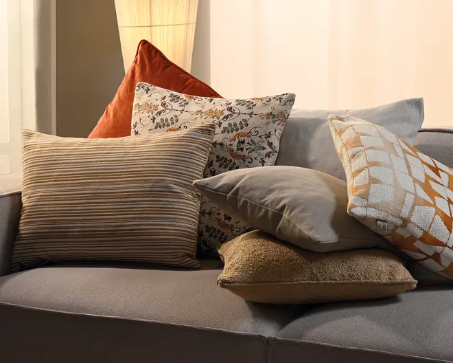En opbygget samling pyntepuder på en grå sofa med beige og brune stribede puder, en pude med blomstermønster, en rustfarvet trekantet pude, en ensfarvet grå pude, en tekstureret tan-farvet pude og en hvid pude med orange geometriske detaljer.