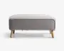 Rektangulær puf med lysegråt tekstilbetræk, afrundede kanter og piping langs toppen. Puffen står på fire skråtstillede ben i lyst træ, hvilket giver et moderne og minimalistisk udtryk.