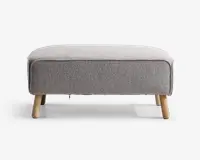 Rektangulær puf med lysegråt tekstilbetræk, afrundede kanter og piping langs toppen. Puffen står på fire skråtstillede ben i lyst træ, hvilket giver et moderne og minimalistisk udtryk.