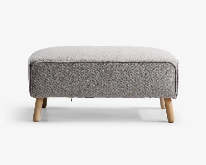 Rektangulær puf med lysegråt tekstilbetræk, afrundede kanter og piping langs toppen. Puffen står på fire skråtstillede ben i lyst træ, hvilket giver et moderne og minimalistisk udtryk.