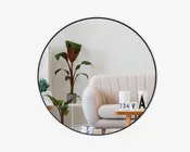 Et rundt spejl med en tynd sort ramme reflekterer en moderne stue. Rummet har en lys beige, polstret sofa med buede armlæn, et sofabord i træ og dekorative genstande, herunder en hvid vase, en kalenderblok og et bogstav 'A' ornament. Der er også grønne potteplanter, som tilføjer et naturligt præg til det minimalistiske interiør.